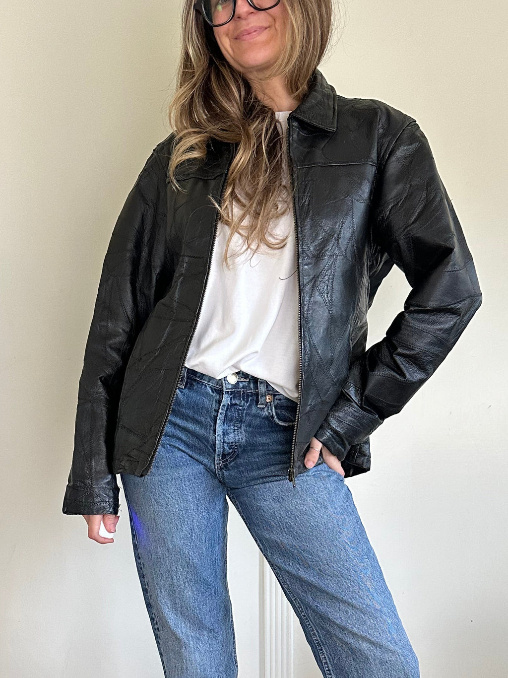 Person wearing a black jacket, white shirt, blue jeans, and black shoes in a room with wooden floor and white walls.