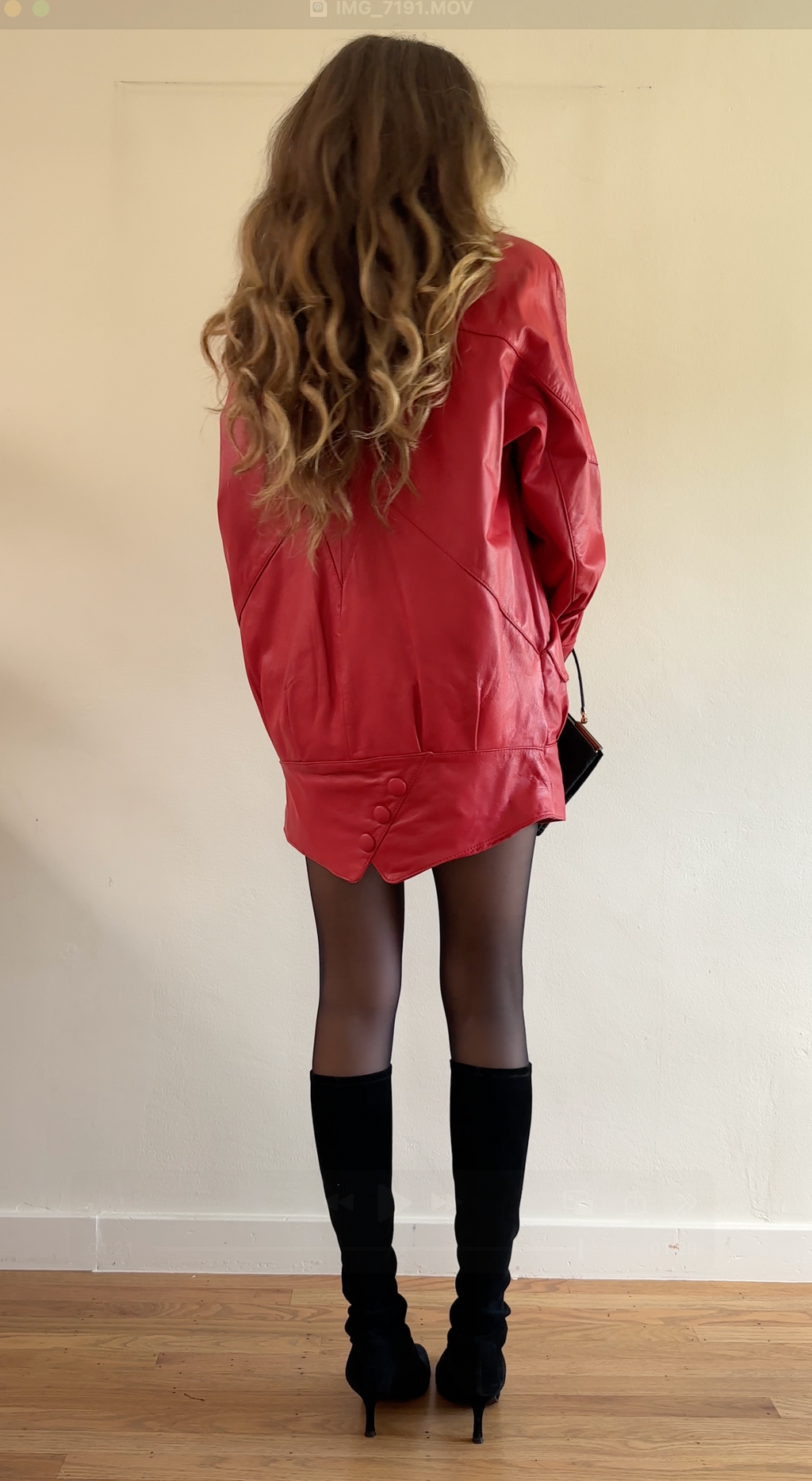 Person wearing a red dress and black knee-high boots standing against a plain wall.